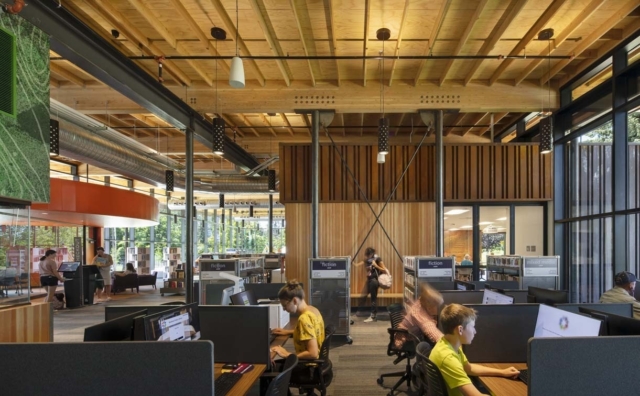 Spacious modern library with exposed wooden beams and tall windows, people at computer workstations, fiction shelves and lounge areas.