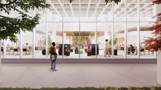 Person with backpack stands outside glass wall watching a bright library atrium where people study at desks and lounge on green seating.
