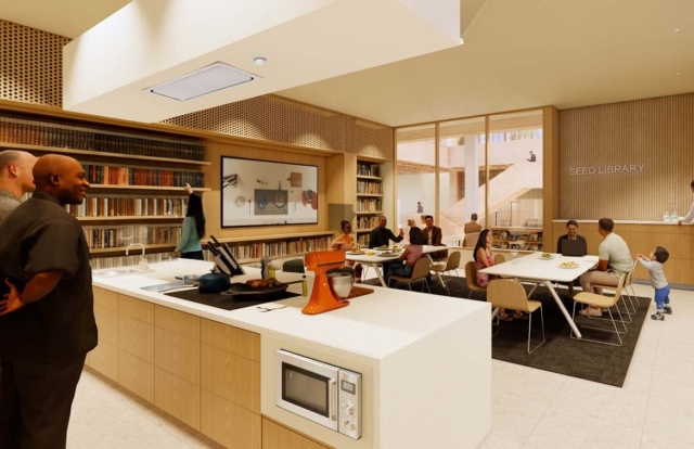 Modern communal kitchen and Seed Library with chefs at an island, orange mixer, microwave, bookshelves and diners conversing at tables.