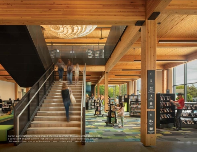 Modern timber library interior with staircase, blurred patrons ascending, patterned carpet, tall windows and patrons browsing shelves.