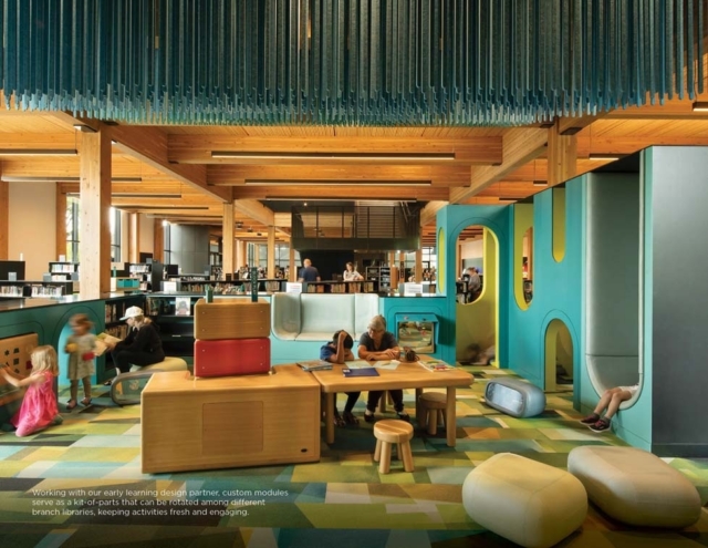 Bright children's library space with exposed wooden beams, colorful modular play nooks, low tables and stools, kids reading and playing.