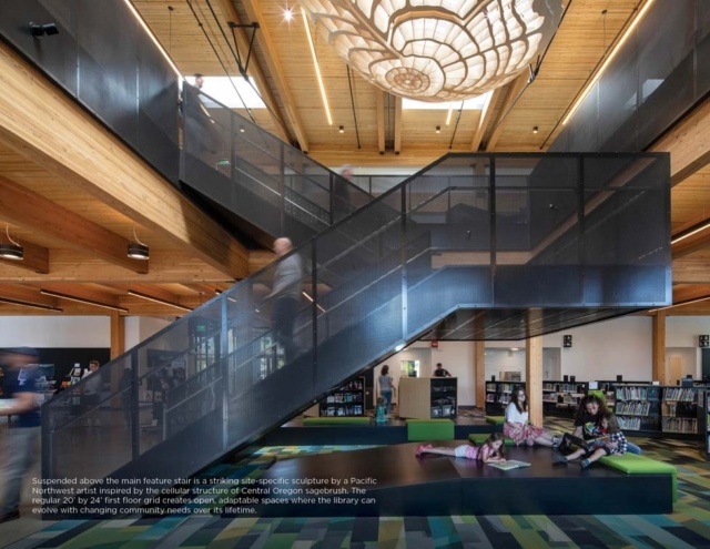 Modern library interior with a large mesh staircase under a wooden ceiling, sculptural pendant light above, children reading on a platform.