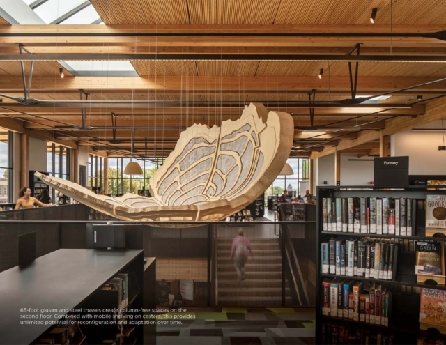 Sunlit modern library with exposed wooden beams and skylights, rows of books and a large suspended wooden leaf sculpture over central stair.