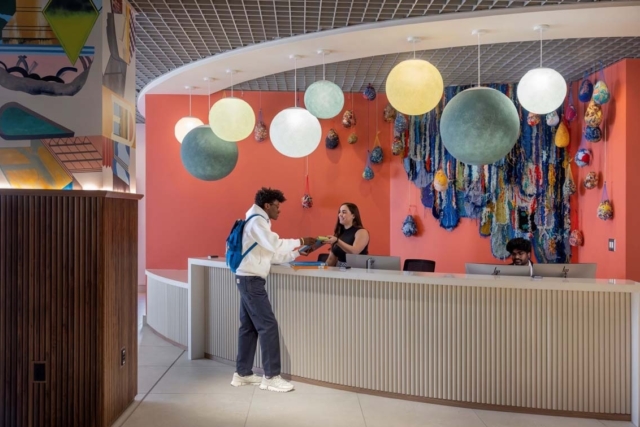 Student with blue backpack hands documents to a smiling staff at a curved desk under globe pendant lights and colorful textile wall art.