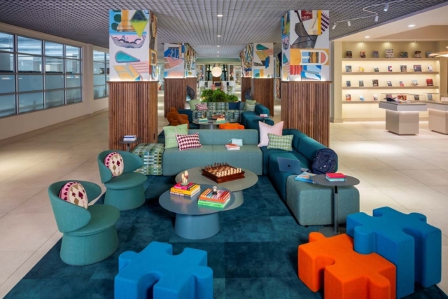 Bright modern lounge with teal sectionals, round tables holding a chessboard and books, puzzle ottomans and patterned mural pillars.