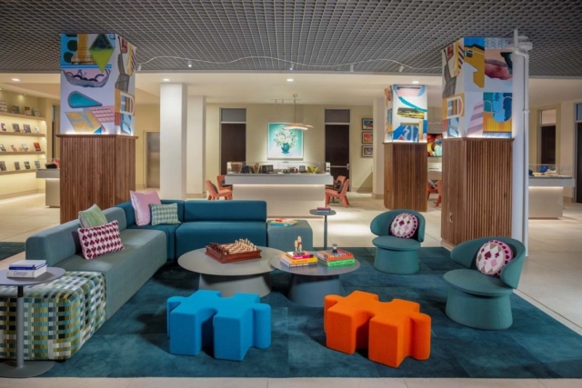 Hotel lounge with teal sofa, patterned cushions, blue and orange puzzle stools, round tables topped with books and a chessboard.