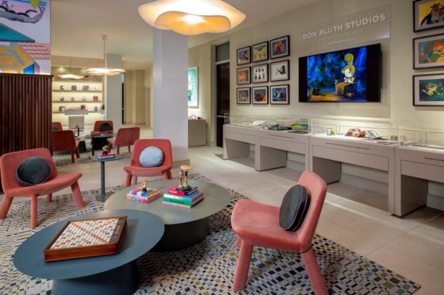 Don Bluth gallery lounge with framed animation and TV, coral chairs with round cushions, and low tables with books and a Scrabble board.