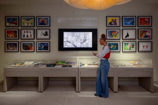 Young woman studies framed Don Bluth animation art and display cases in a gallery, a central screen shows a black-and-white storyboard.