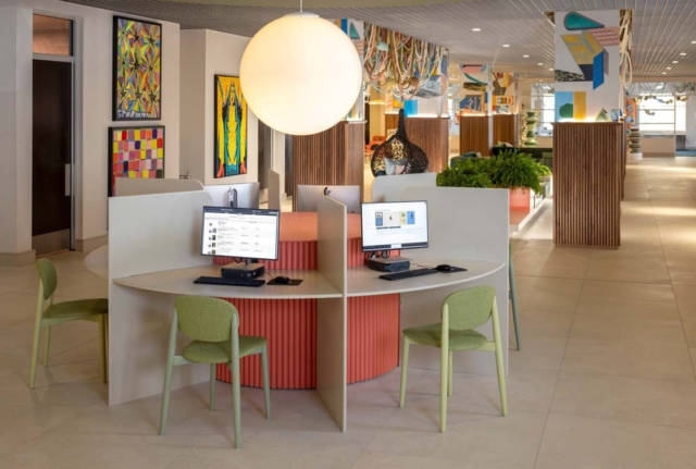 Modern hotel business center with a round four-station desk, dual monitors, mint green chairs, large globe light and colorful wall art.