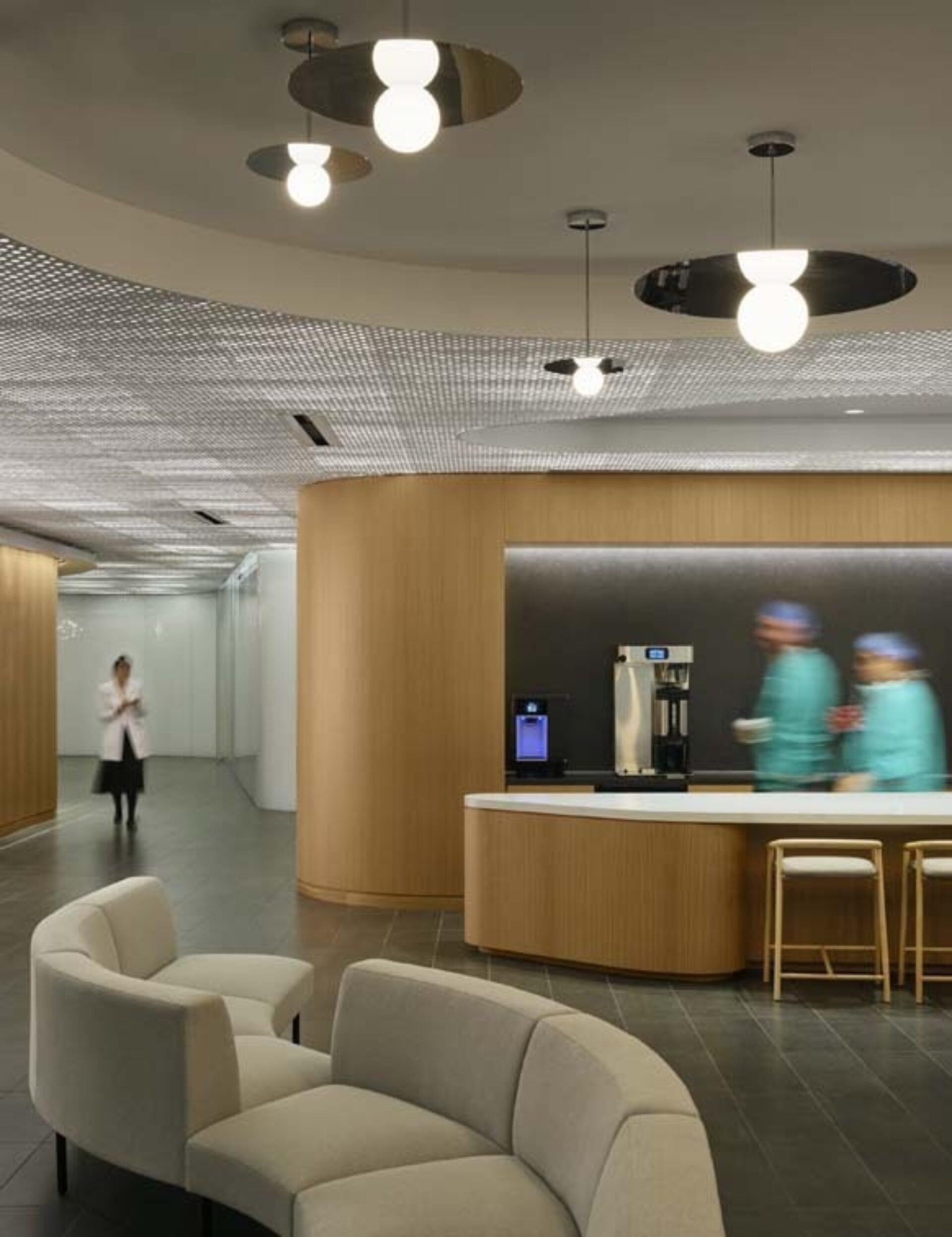 Stylish hospital lounge with curved wood coffee bar, globe pendant lights, beige sofas, blurred green-scrub staff and a clinician.