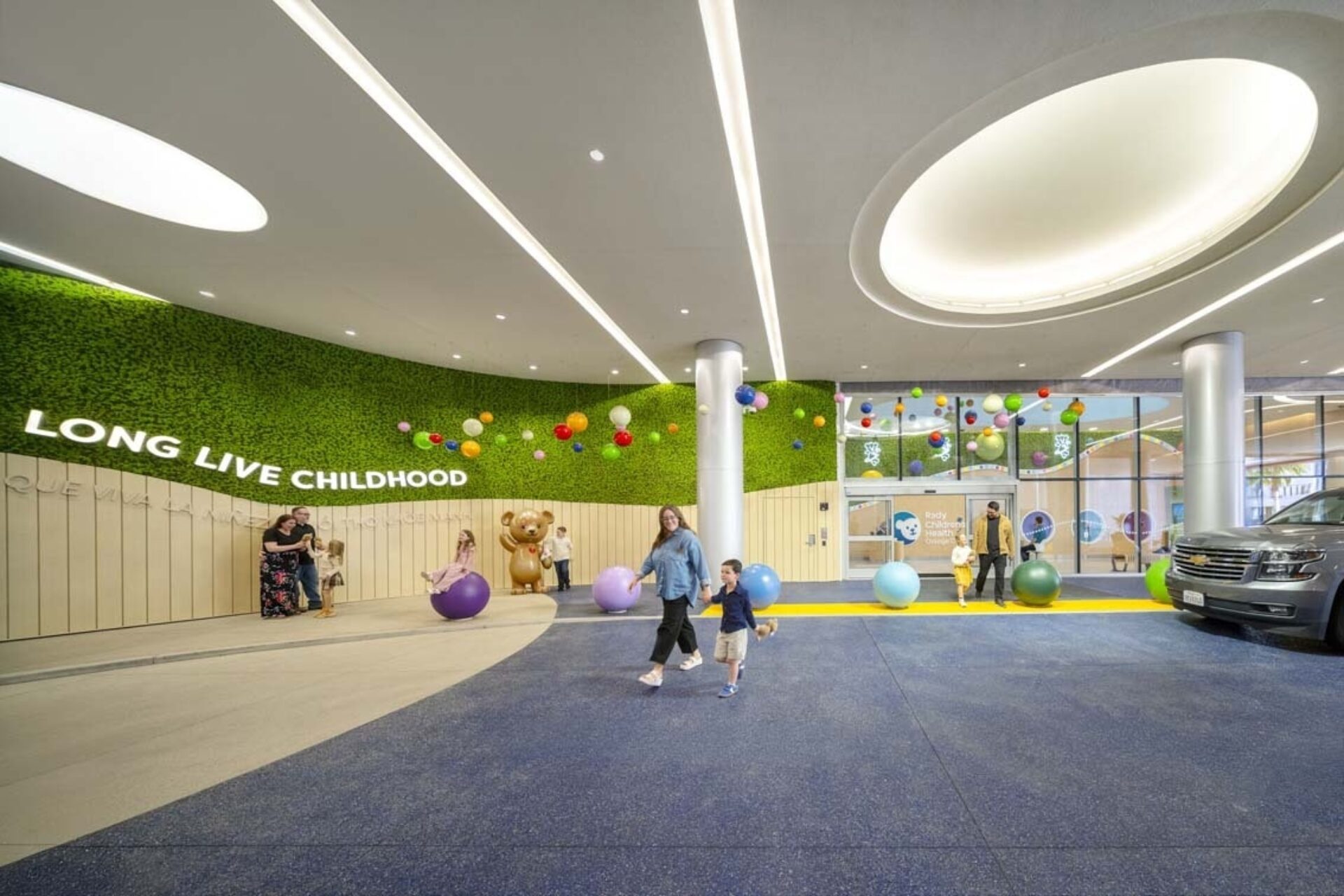 Pediatric entry with green wall reading 'LONG LIVE CHILDHOOD', colorful hanging spheres, kids playing on oversized bouncy balls.