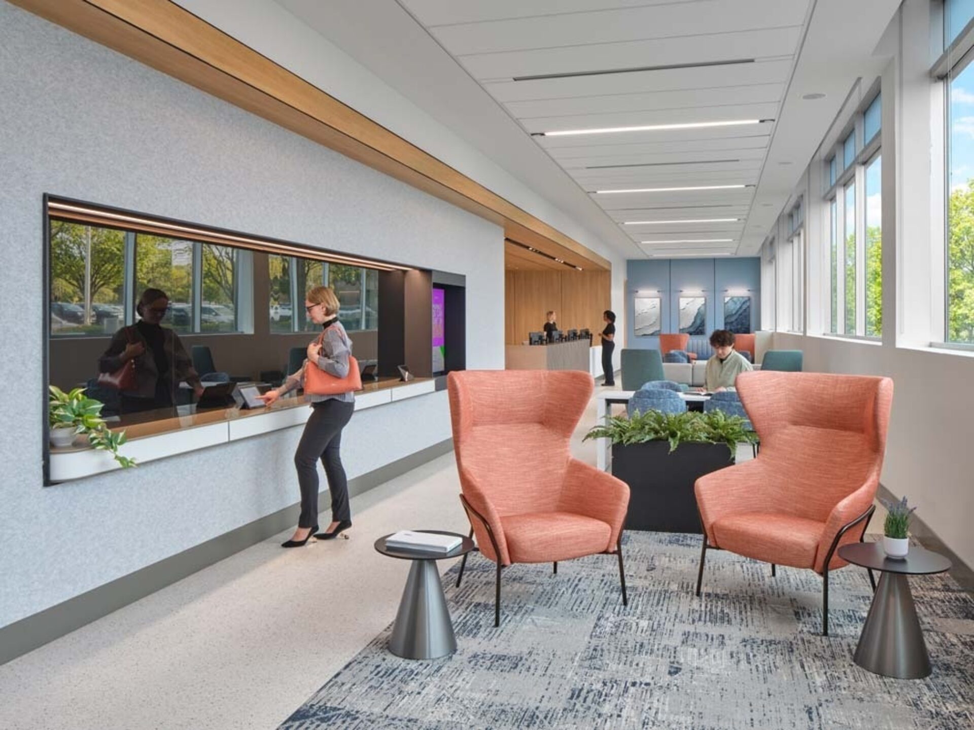 Bright modern office lounge with two coral wingback chairs facing a planter, small side tables, a long service window and people at desks.
