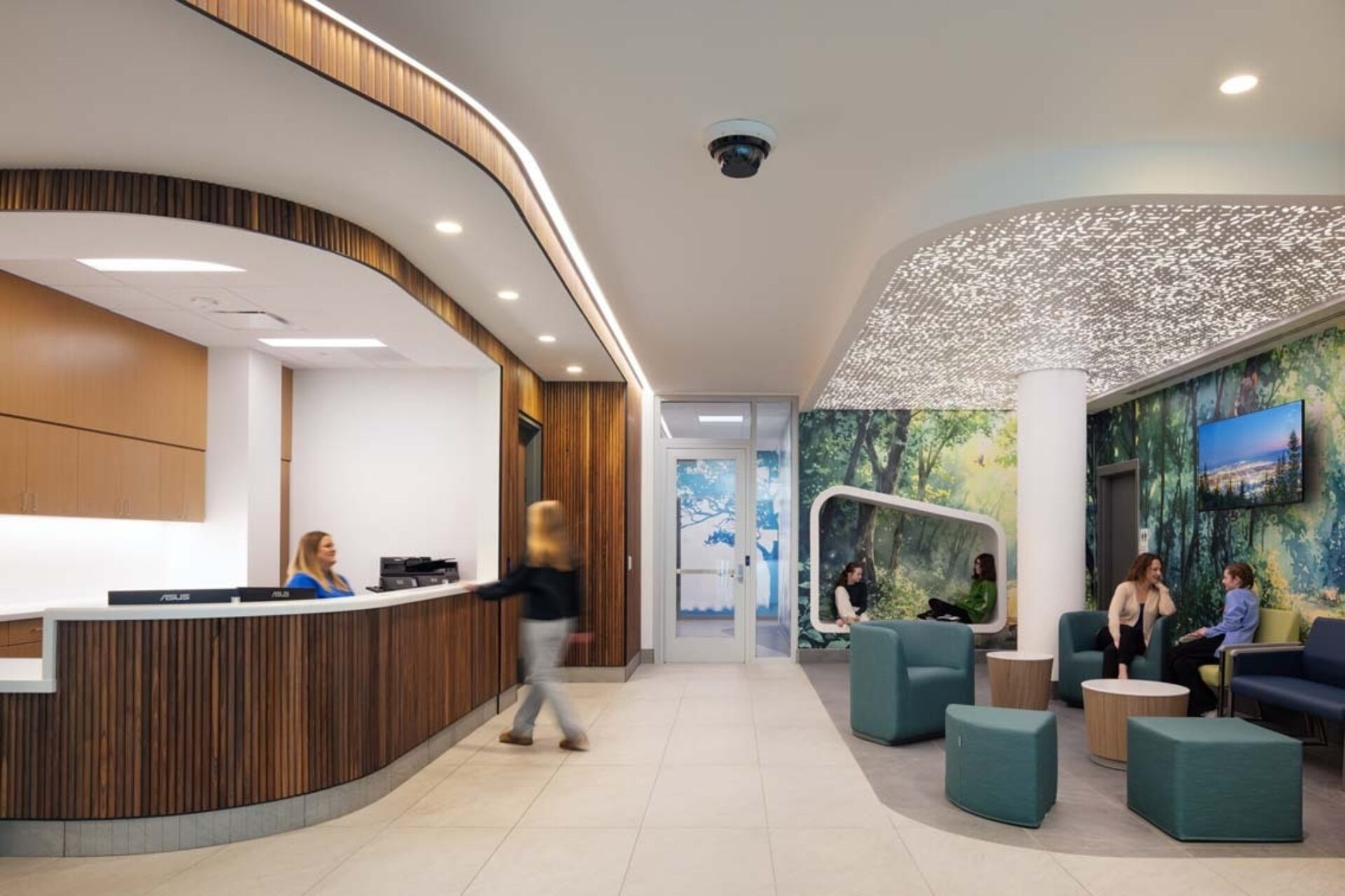 Modern reception lobby with curved wood desk, blurred person walking, green lounge seating, forest mural and perforated illuminated ceiling.