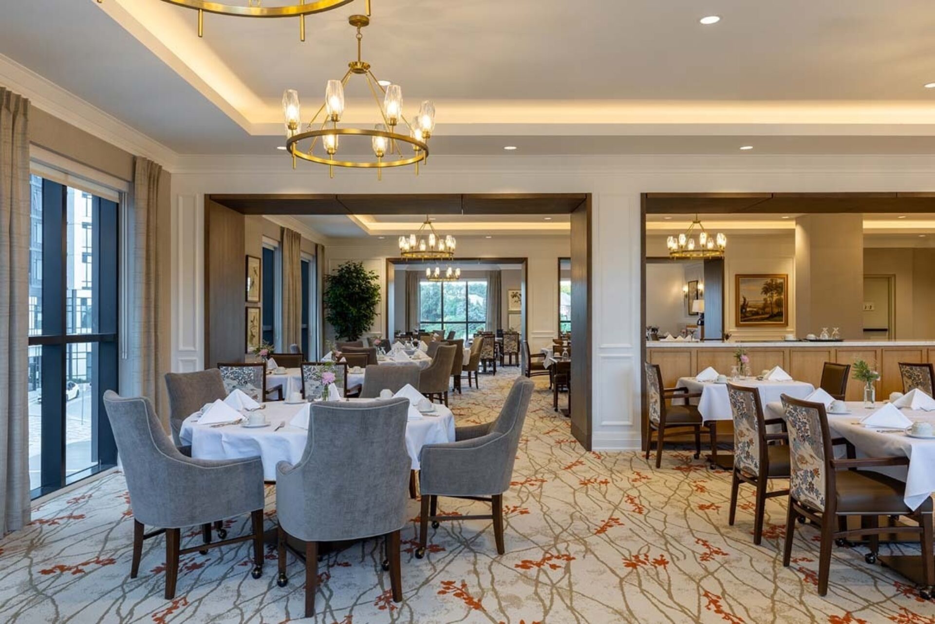 Elegant dining room with round tables draped in white linens, gray chairs, brass chandeliers, floral centerpieces and patterned carpet.