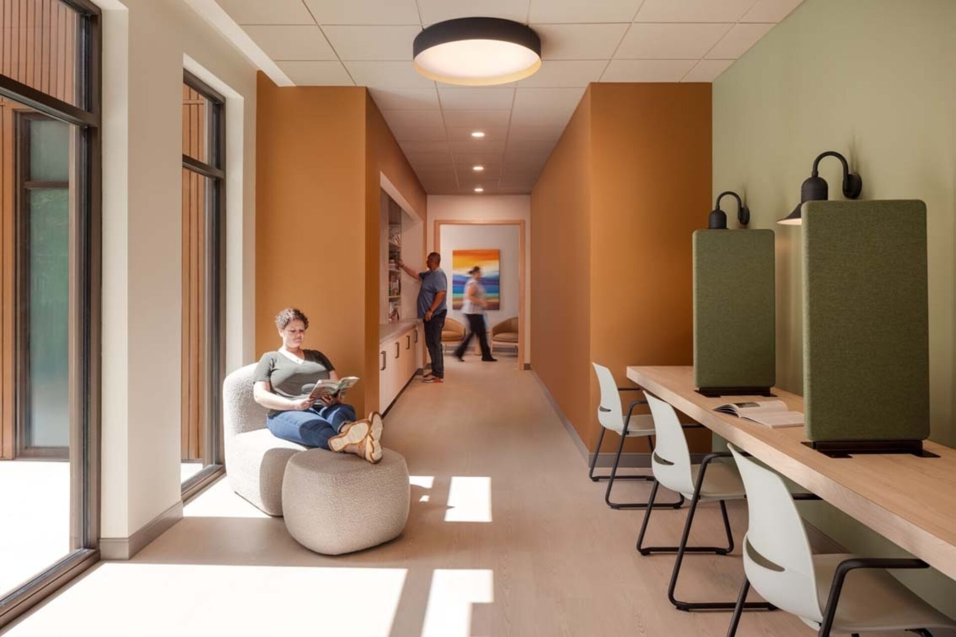 Sunlit waiting corridor with a woman reading on a cushioned chaise by tall windows, study carrels and people near a reception area.