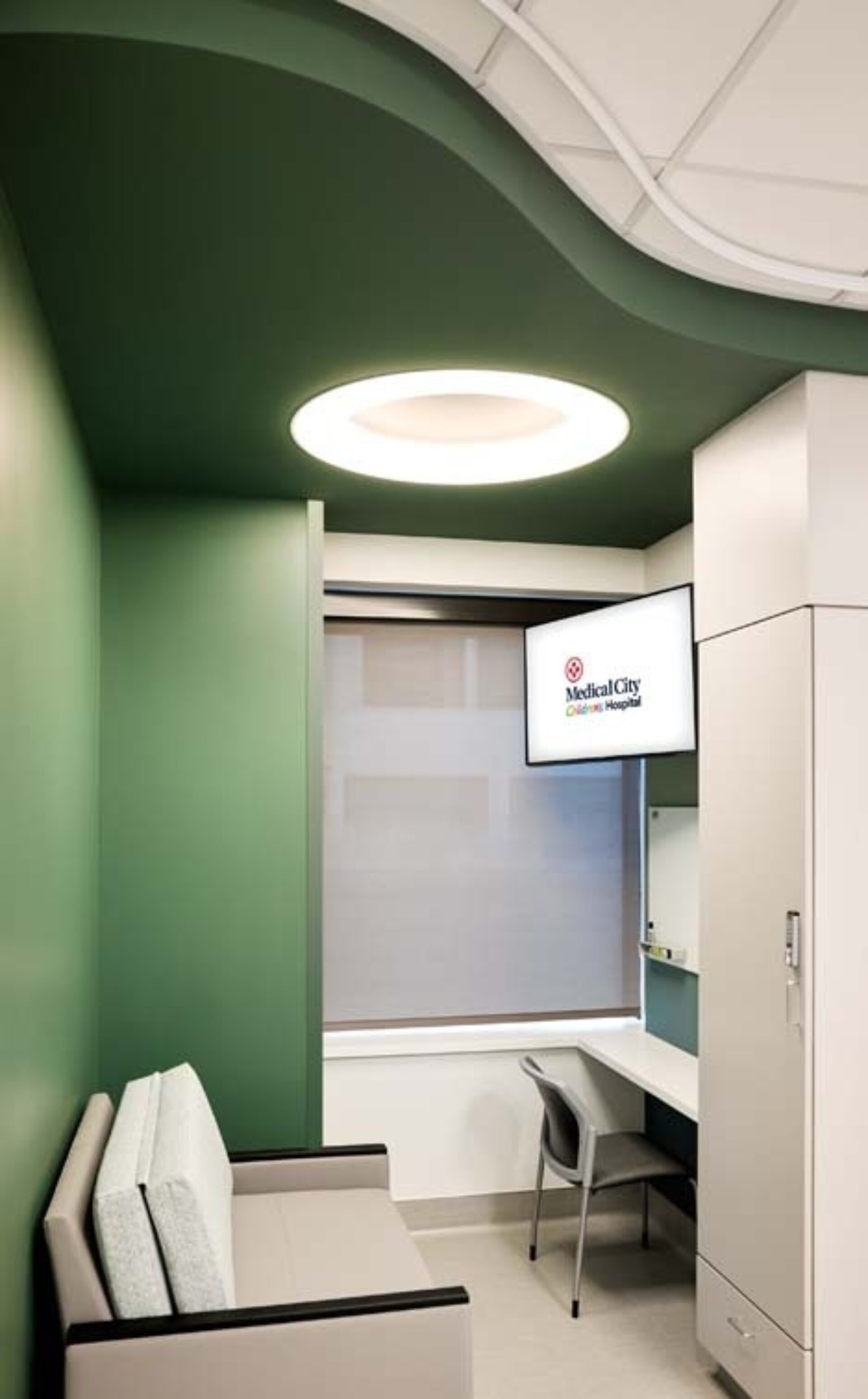 Small clinical room with green curved ceiling, round recessed light, gray bench, desk and chair, TV showing Medical City Clinic & Hospital.