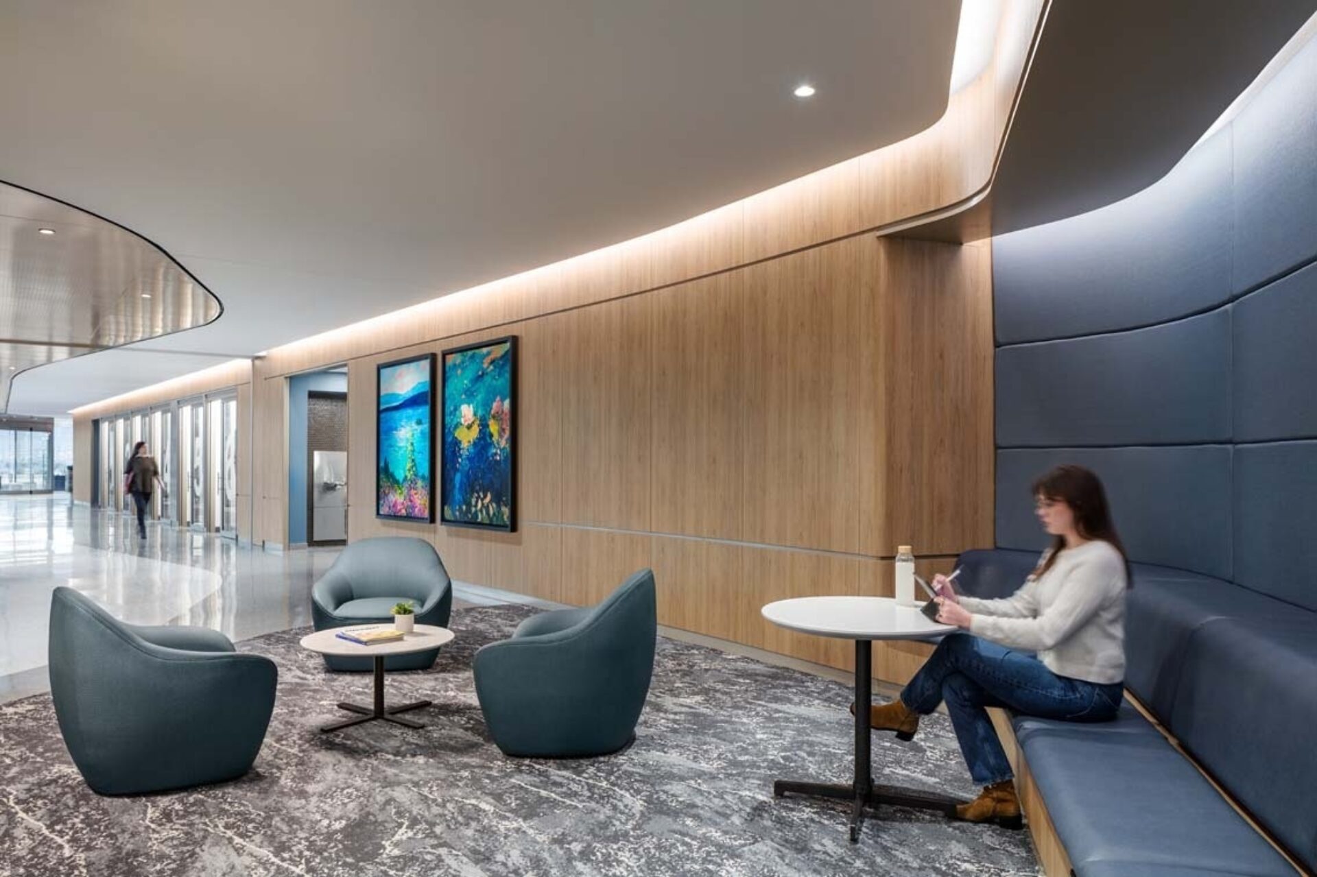 Modern office lounge with curved wood wall, two vibrant framed paintings, three teal chairs, low round table, and a woman using a tablet.