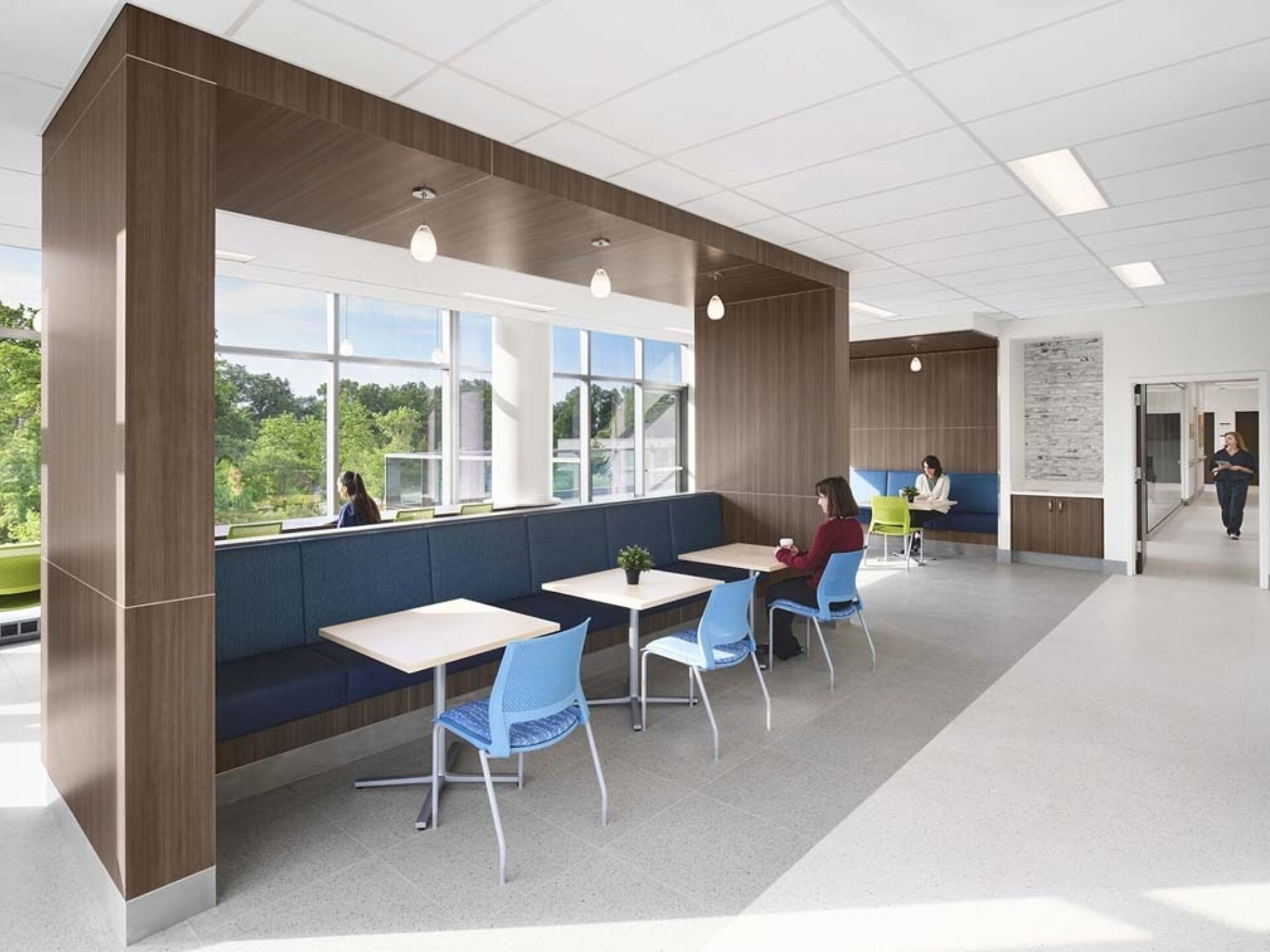 Modern hospital waiting area with wood-paneled booths, navy banquettes, light-blue chairs, small tables, large windows and people seated.