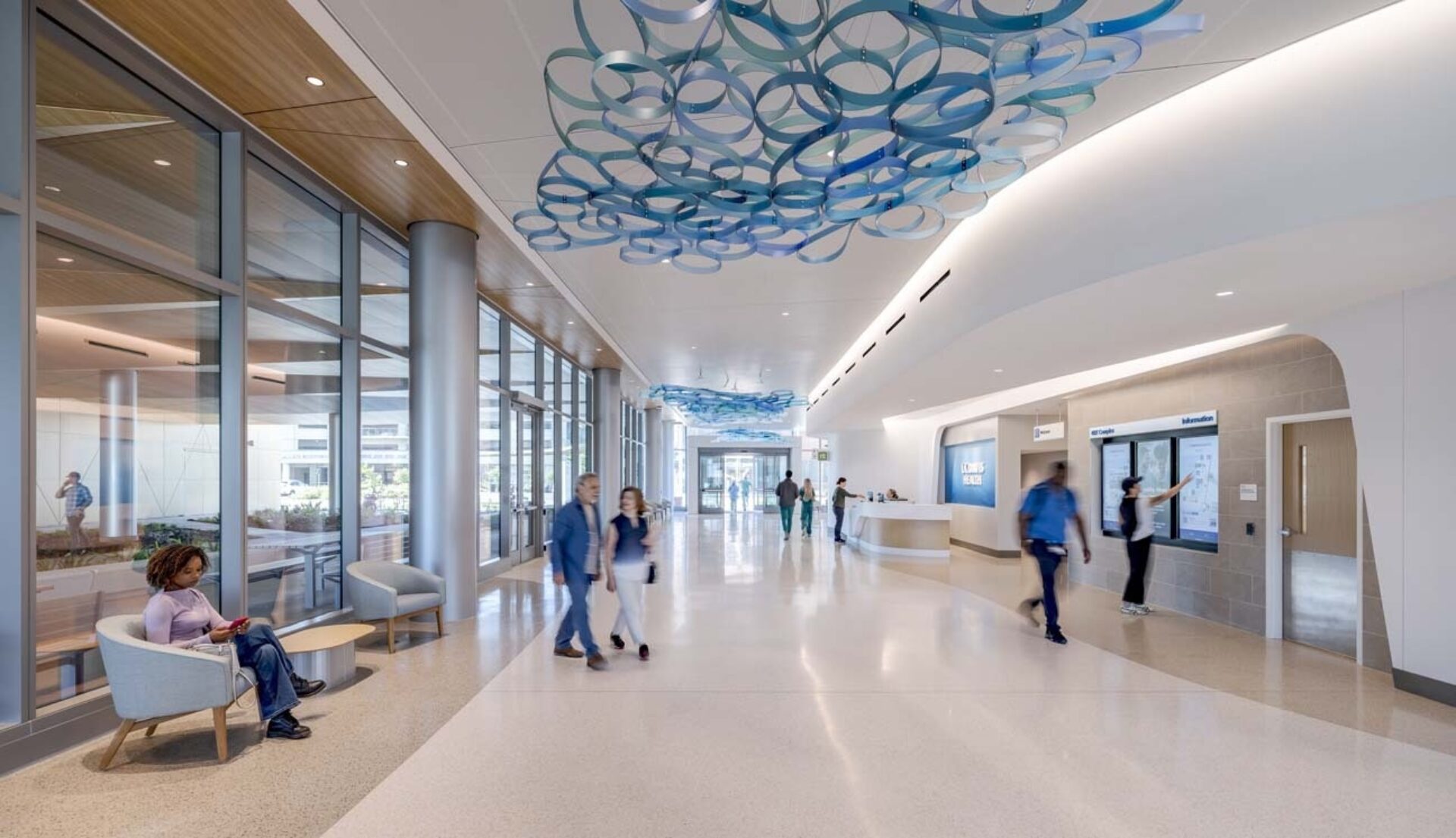 Bright hospital lobby with suspended blue circular ceiling sculpture, glass wall and seating left, reception desk and directory right.
