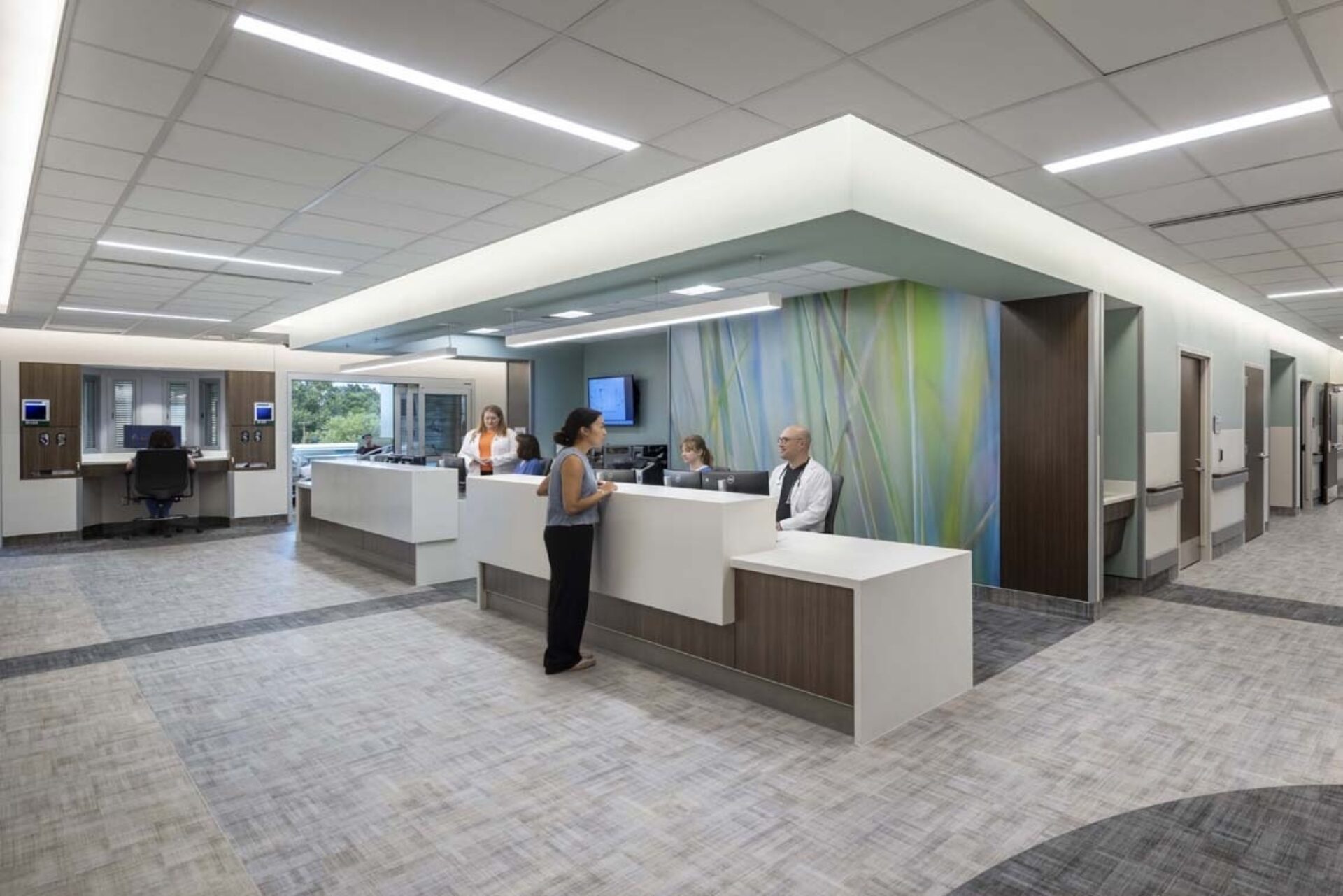Bright, modern hospital nurses' station with reception desks, staff helping a visitor, abstract green-blue wall art, wide carpeted corridor.