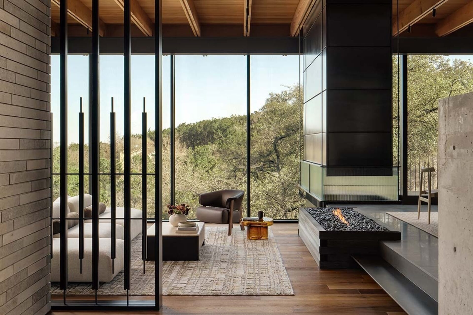 A modern living space with large windows, showcasing a cozy seating area, decorative fireplace, and a view of greenery outside.