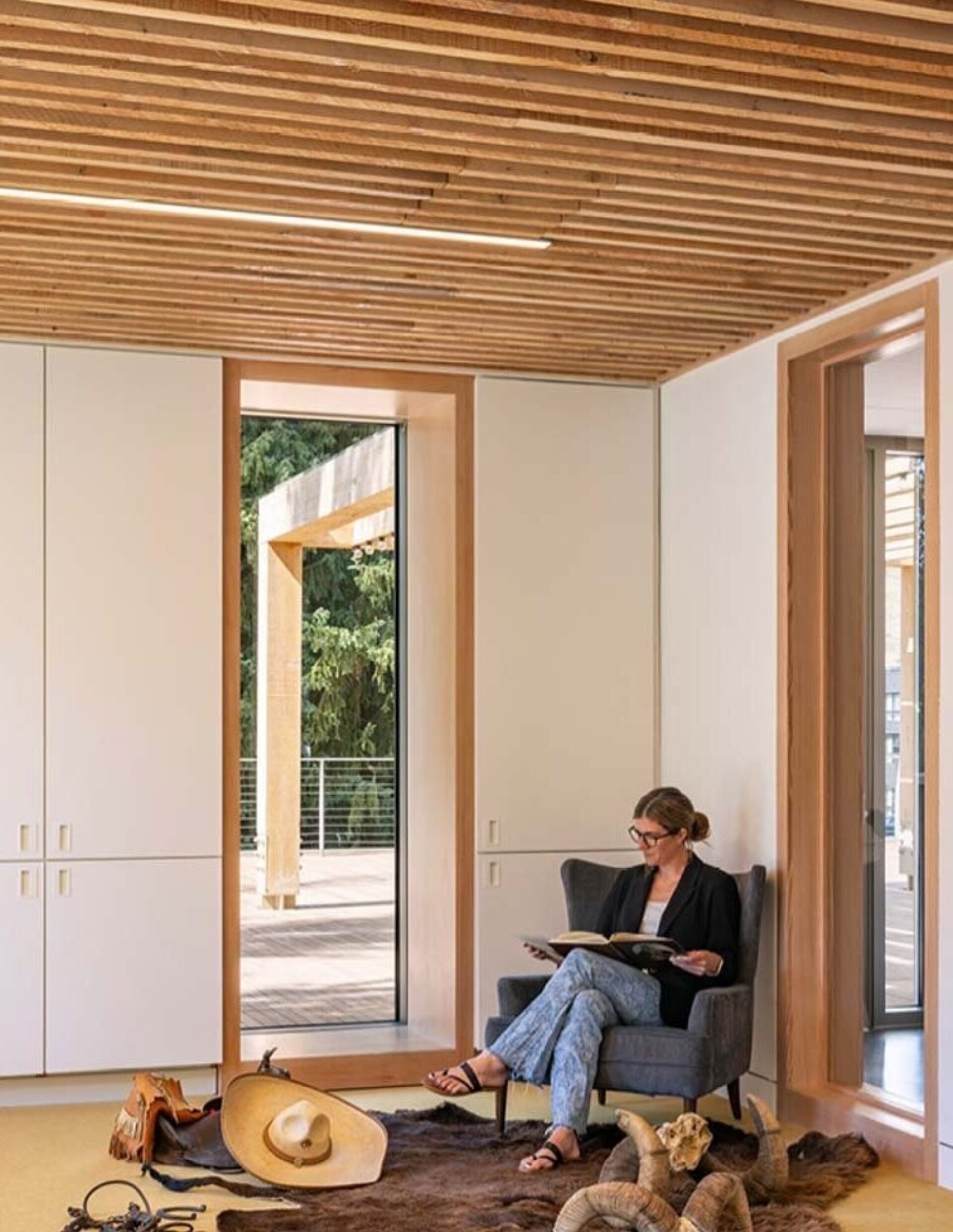 A woman sits in a cozy, modern room with wooden accents, reading a book near large windows overlooking greenery. Decor includes casual accessories.