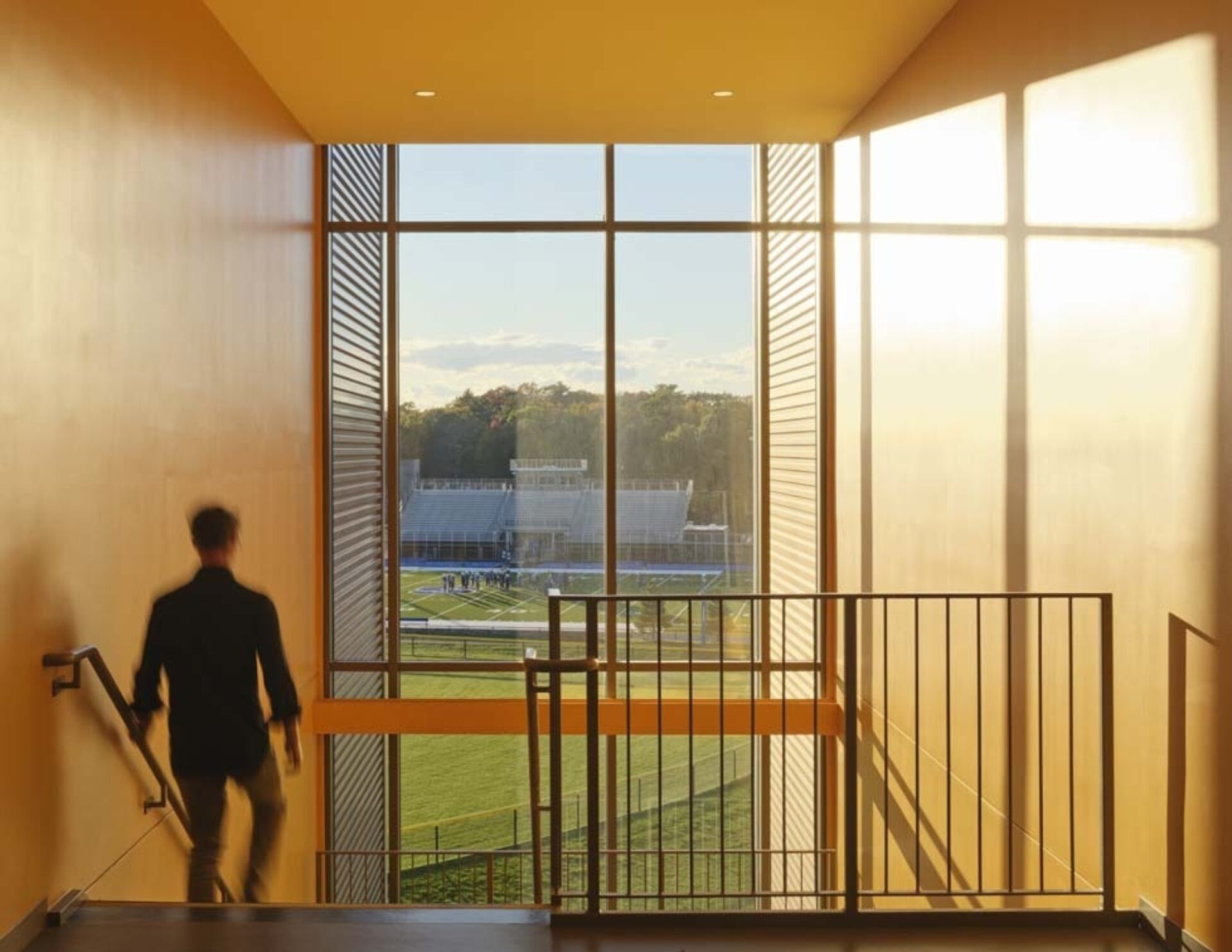 A person descends a staircase framed by large windows, revealing a vibrant view of a sports field bathed in golden sunlight.
