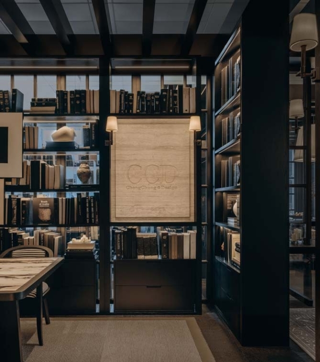 A cozy library corner features dark wooden shelves filled with books and decorative objects, illuminated by stylish wall sconces.