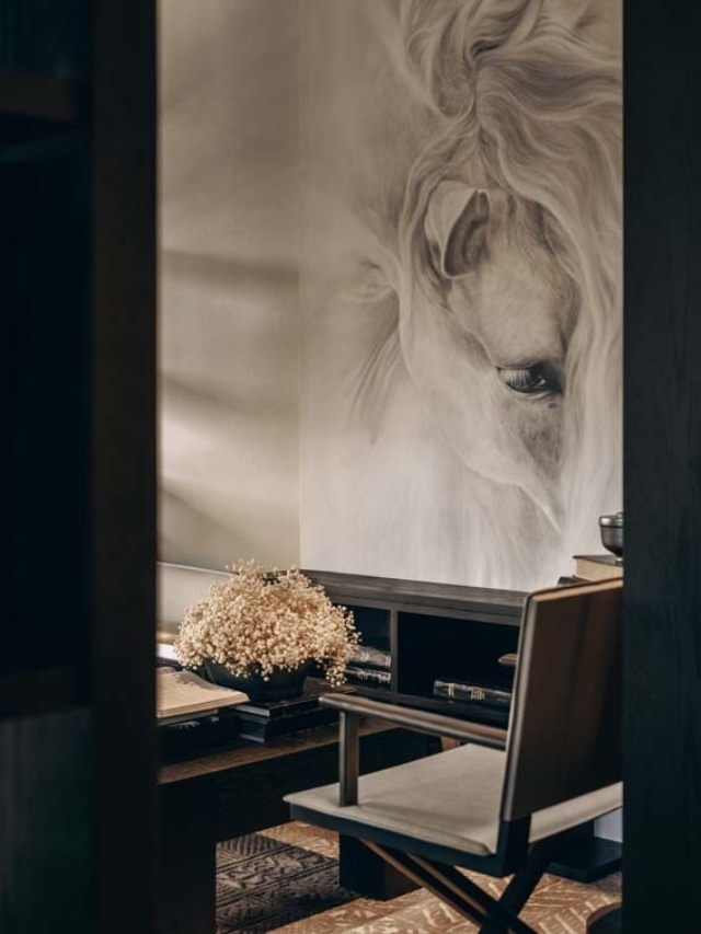 A softly lit room features a detailed horse mural, a wooden desk with books, and a chair next to a floral arrangement in a vase.