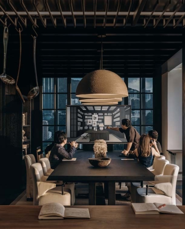 A group of people gathers around a large dining table, focused on a presentation screen showing a design layout in a stylish conference room.