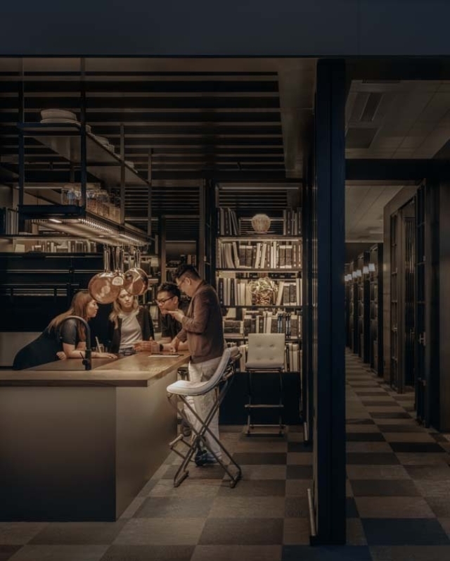 A cozy bar scene features five people engaged in conversation around a large countertop, surrounded by shelves filled with books.