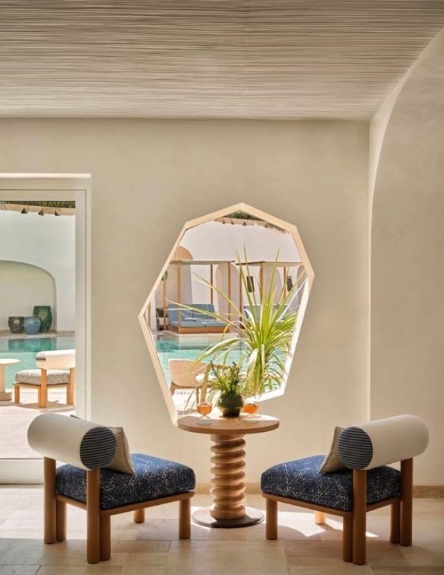 Two stylish chairs with blue cushions and white armrests flank a small table with a plant, framed by a hexagonal window overlooking a pool.