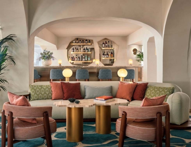 A stylish lounge area features a plush sofa with colorful pillows, a sculptural table, and cozy chairs, set against a bar backdrop.