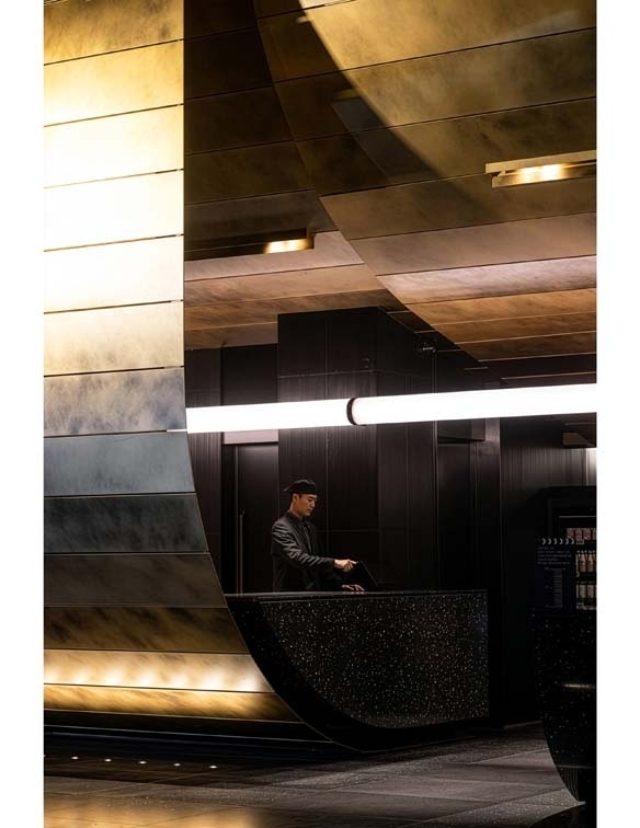 A stylish lobby with modern architecture features a staff member in a black outfit at a sleek reception desk under soft lighting.