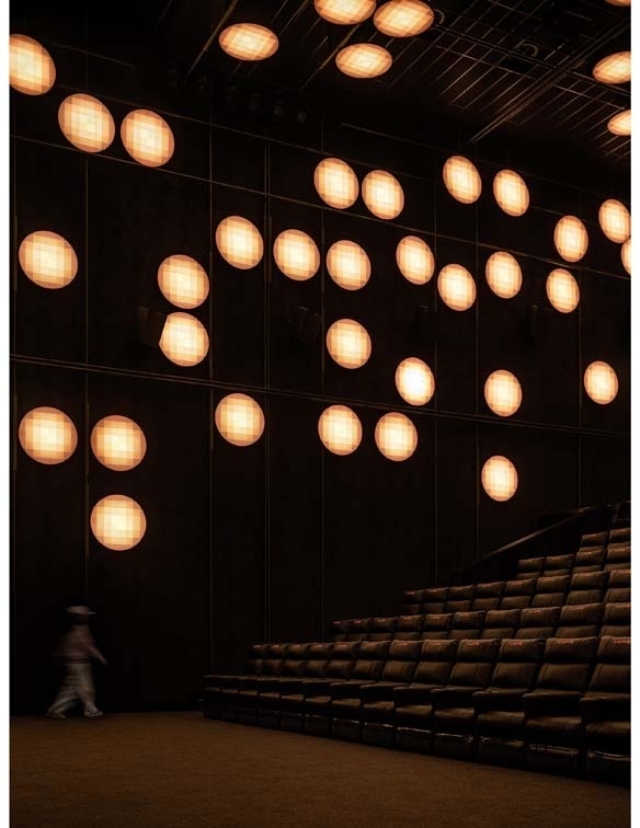 A modern auditorium features a dark wall illuminated by various circular light fixtures, while rows of empty brown seats provide seating.