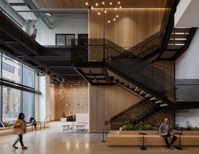 A modern building lobby features sleek staircases, wooden accents, contemporary lighting, and people engaging in casual conversation.