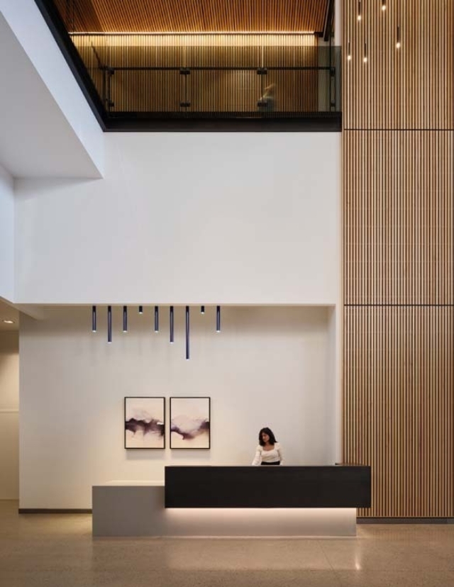 A modern reception area features a sleek desk, a woman working behind it, and artistic wall decor with wooden paneling above.