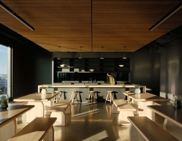 A modern dining area features light wood tables and benches, with a sleek kitchen backdrop and a person preparing food under soft lighting.