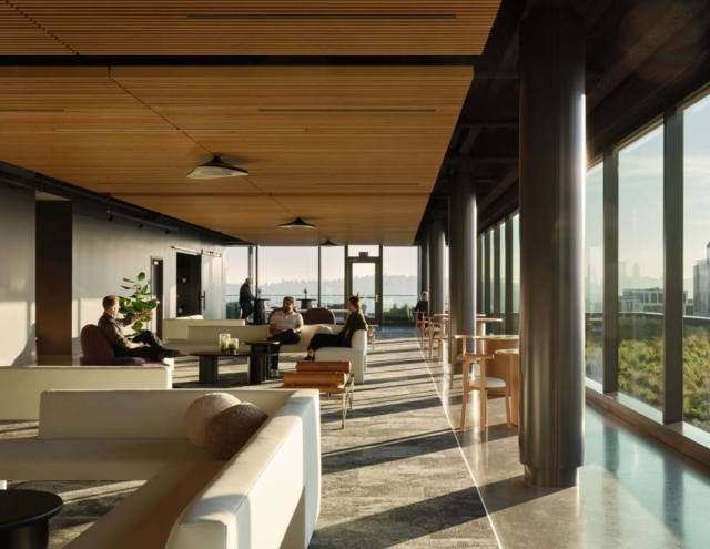 A modern lounge features large windows, natural light, and various seating arrangements where people engage in conversation and work.