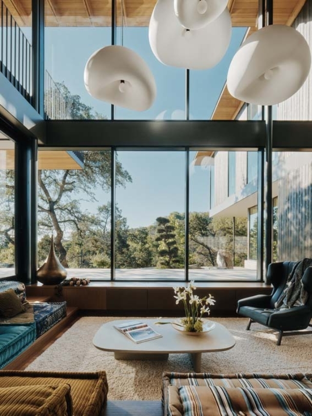 A modern living room features large glass windows, soft furnishings, a stylish coffee table, and decorative lighting hanging above.