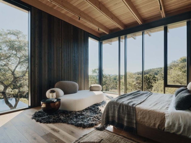A modern, airy bedroom features large windows, a cozy bed, and a stylish lounge chair, surrounded by scenic greenery outside.
