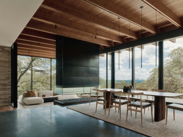 A modern interior features wooden beams, large glass windows with scenic views, a sleek black fireplace, and a minimalist dining area.