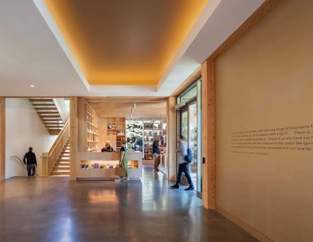 A spacious, modern lobby features wooden accents, a reception area with bookshelves, and visitors engaging in conversation and movement.