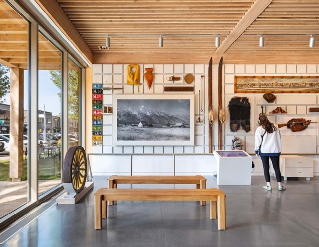 A modern exhibit space features wooden benches, a large black-and-white landscape photo, eclectic historical artifacts, and natural light.
