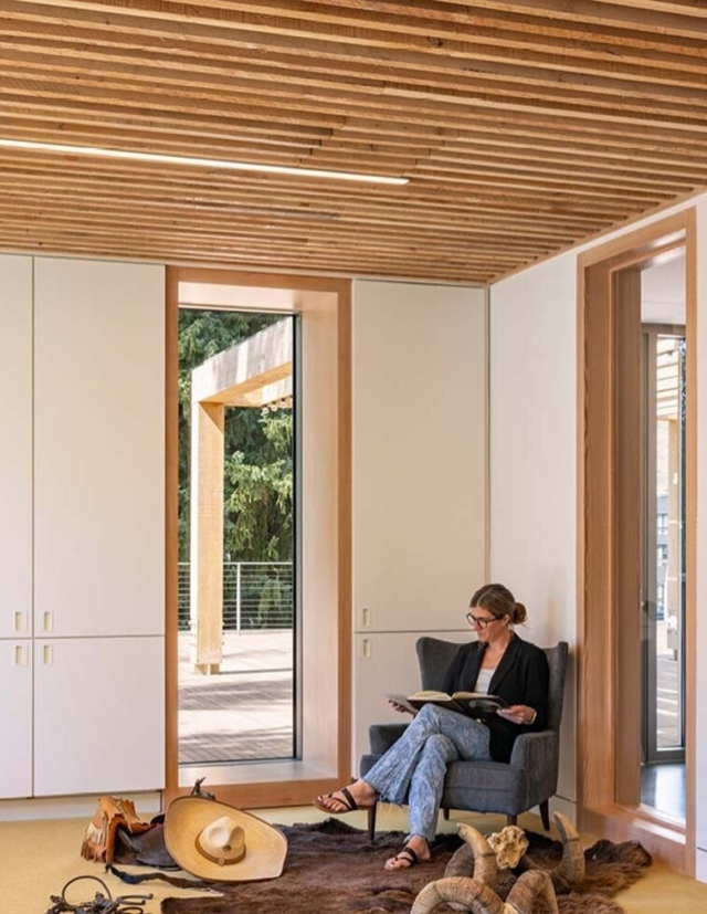 A woman sits in a cozy, modern room with wooden accents, reading a book near large windows overlooking greenery. Decor includes casual accessories.