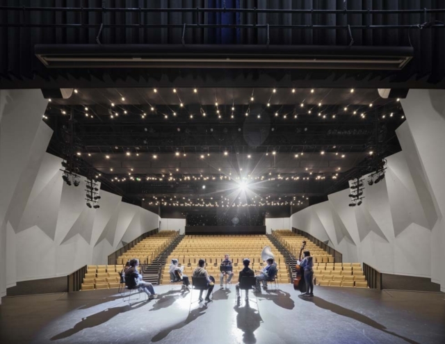 A modern performance space with a circular stage, featuring musicians seated in a semi-circle, and tiered seating in the background.