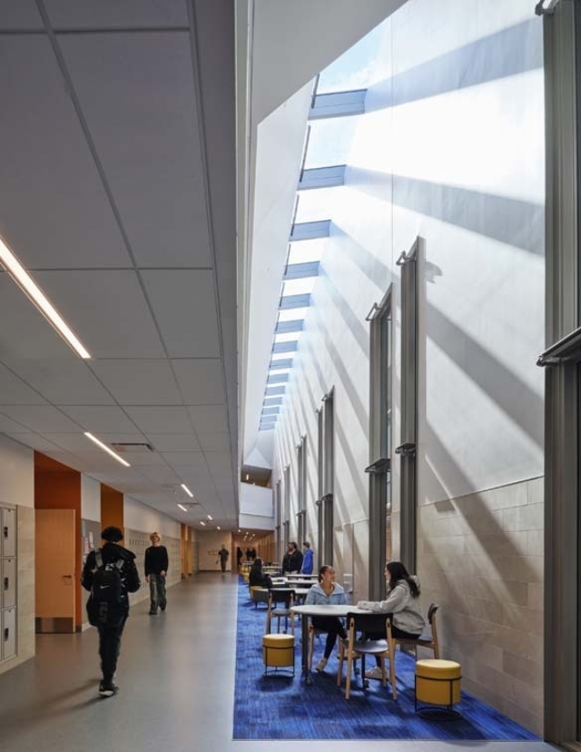 Brightly lit hallway features large windows and skylights, with students sitting at tables and others walking through the space.