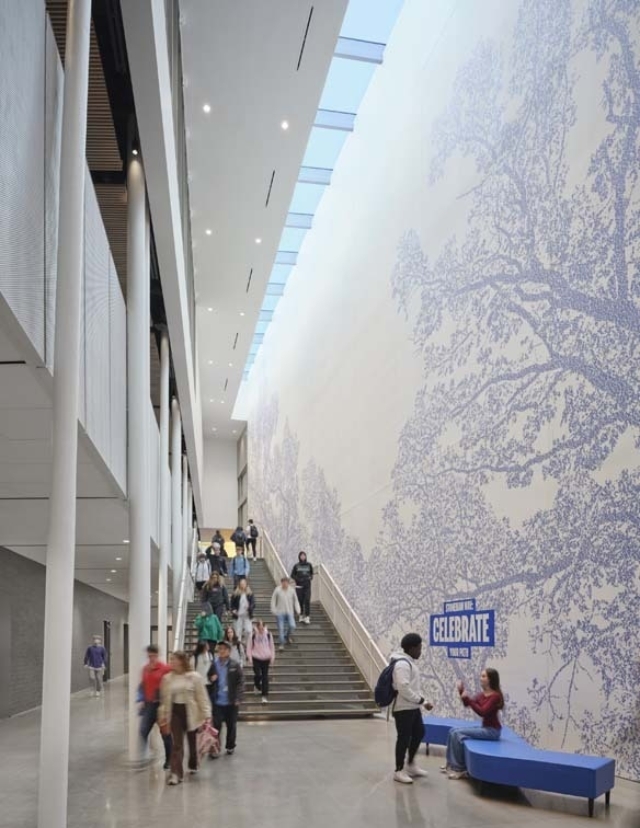 A spacious modern atrium features a large mural of trees, with people walking down the stairs and sitting on a blue bench.