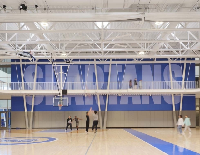 A modern gymnasium featuring a basketball court, with players shooting hoops and a large "SPARTANS" banner displayed prominently above.