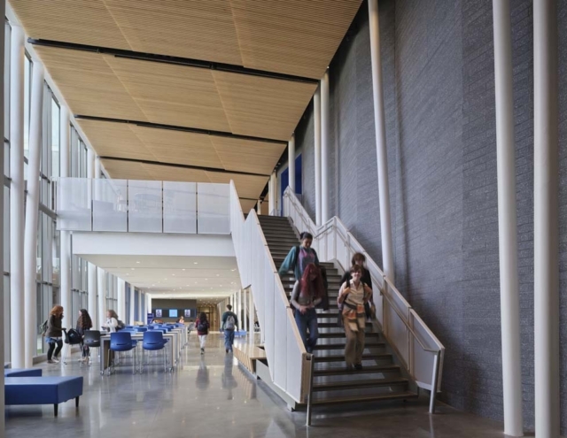 A spacious, modern building interior features large windows, wooden ceilings, and people using the stairs, with tables in the background.